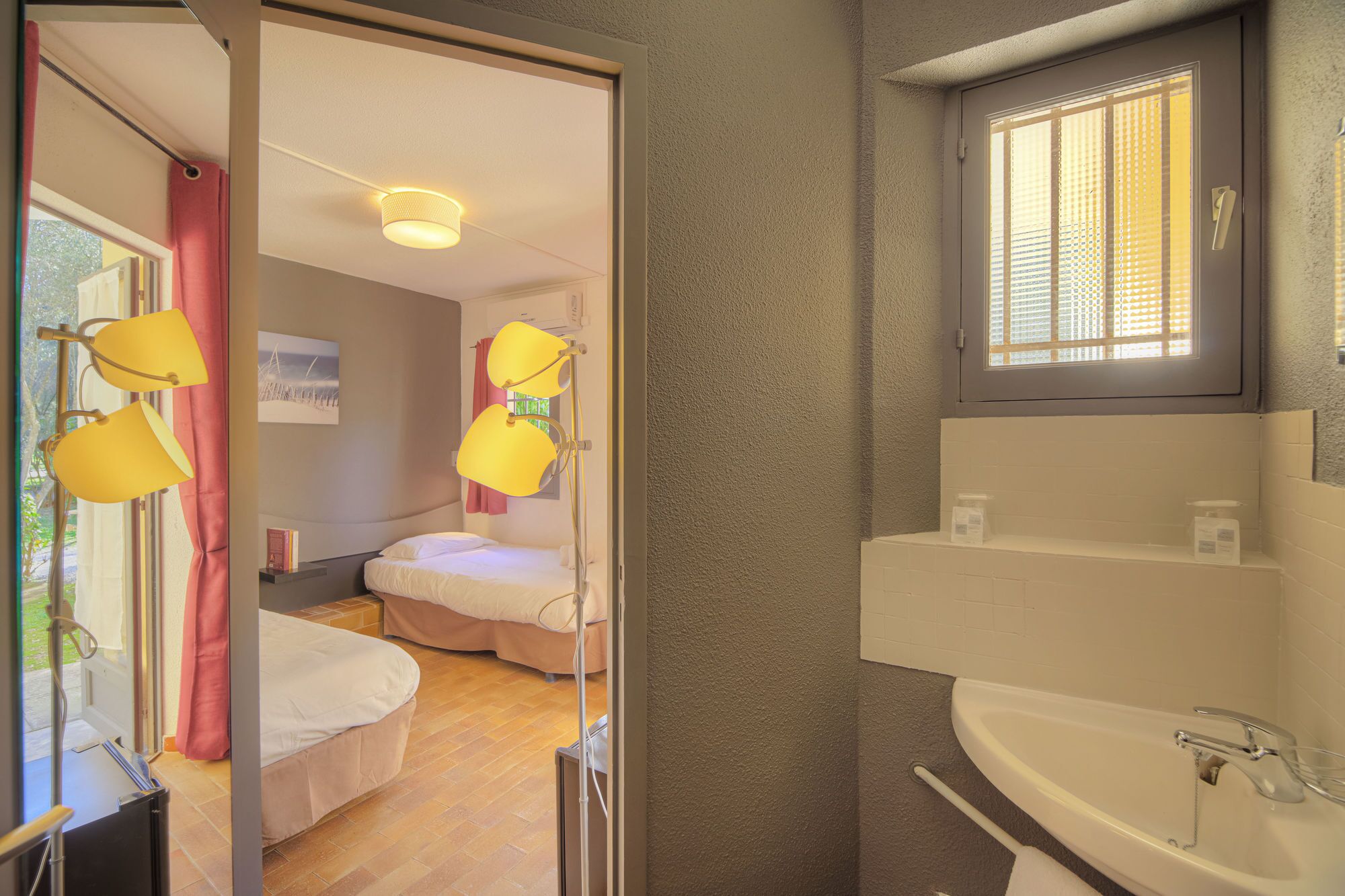 A hotel room with a bed, a bedside lamp, a window with blinds, a bathroom with a sink, and a shower area.