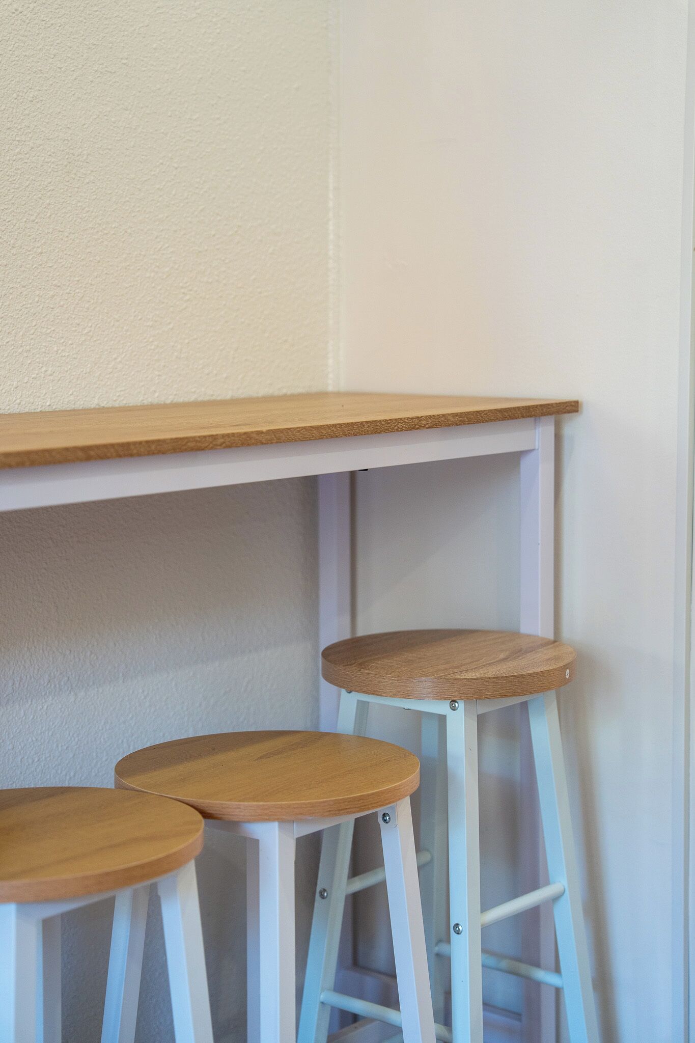 Una esquina con una pared blanca, un estante de madera y tres taburetes blancos con asientos de madera.