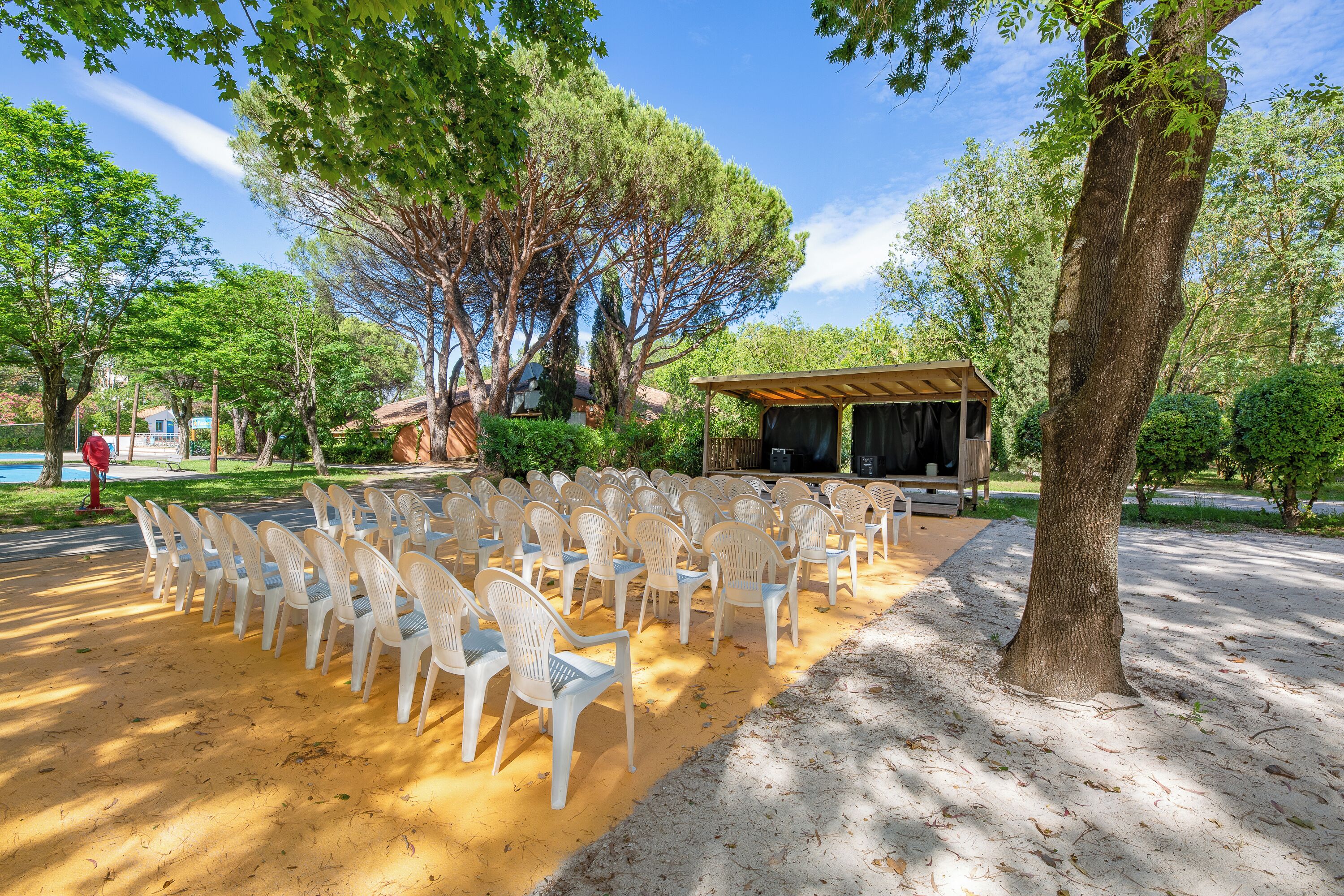 Un espace extérieur aménagé avec des rangées de chaises en plastique blanches disposées sur une surface sablonneuse, entouré d’arbres et d’un pavillon couvert.