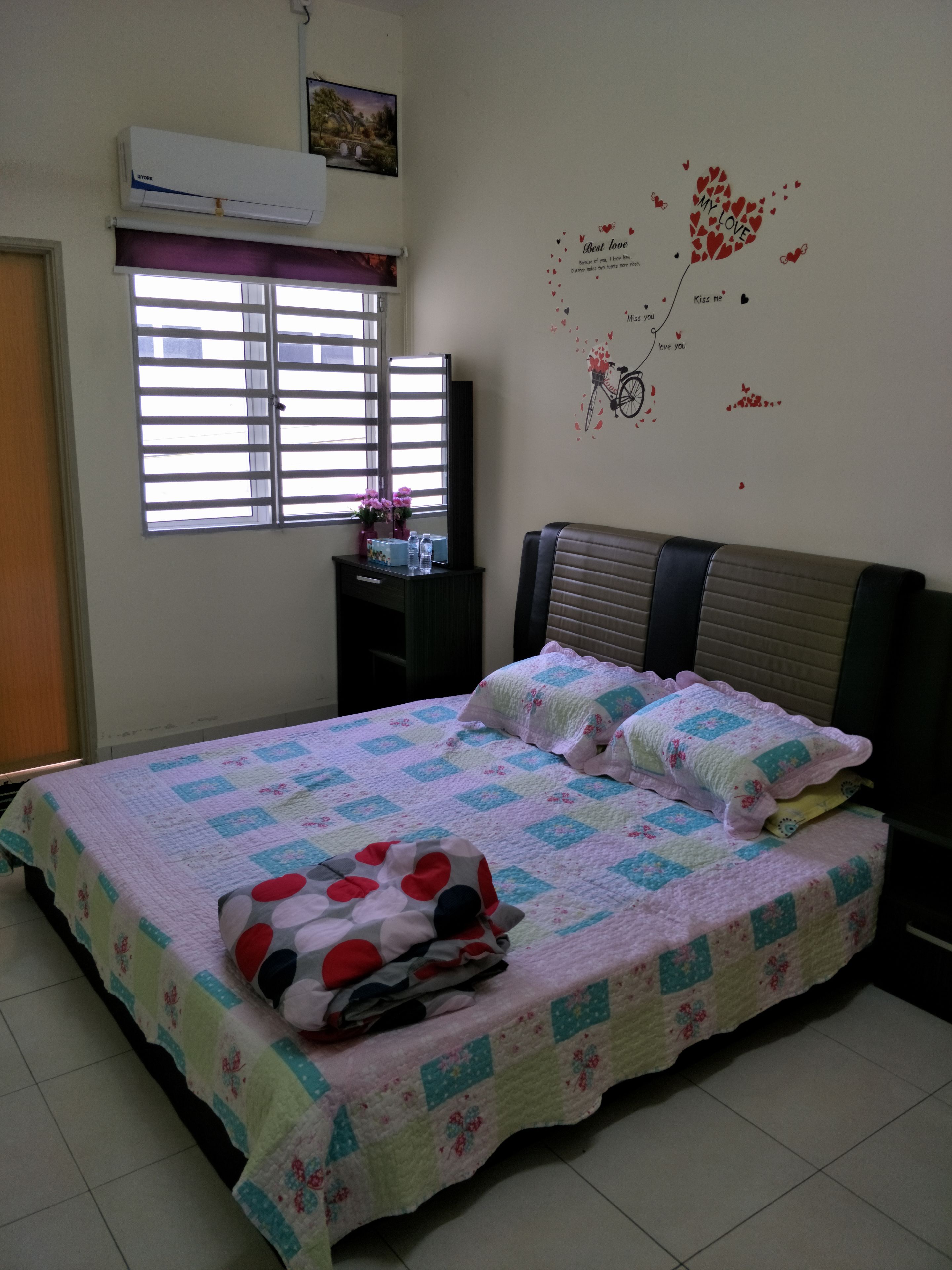A bedroom with a bed, a window with blinds, a wall decoration, and a small table with a vase and bottles.