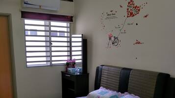 A bedroom with a bed, a window with blinds, a wall decoration, and a small table with a vase and bottles.