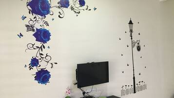 A room with a TV on a stand, a small shelf with a plant, and decorative wall art including blue roses and a street lamp.