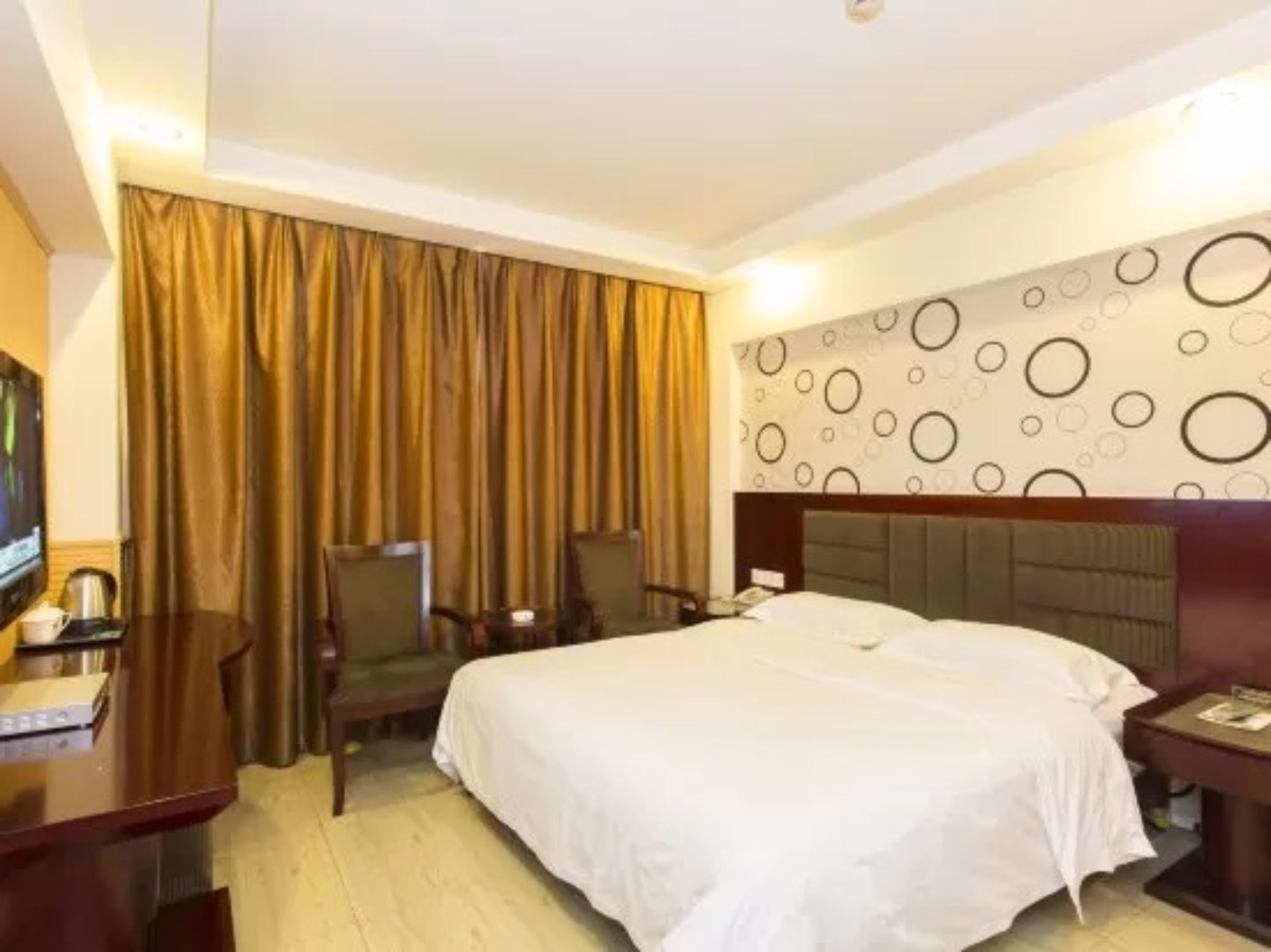 A hotel room with a bed, a desk, a chair, a TV, and a wall with circular patterns.