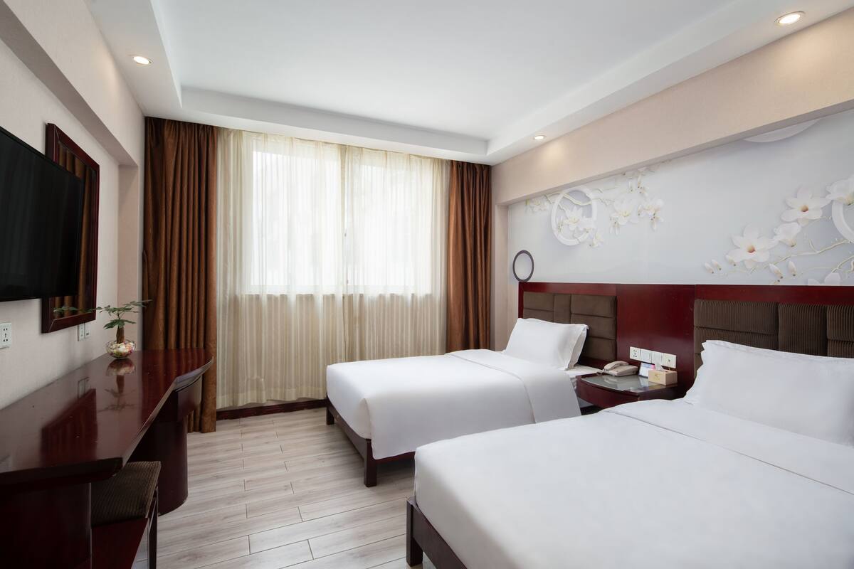 A hotel room with two beds, a desk, a TV, and a window with curtains.