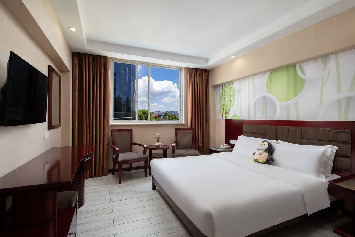 A hotel room with a large bed, two chairs, a small table, a TV, and a window with curtains.