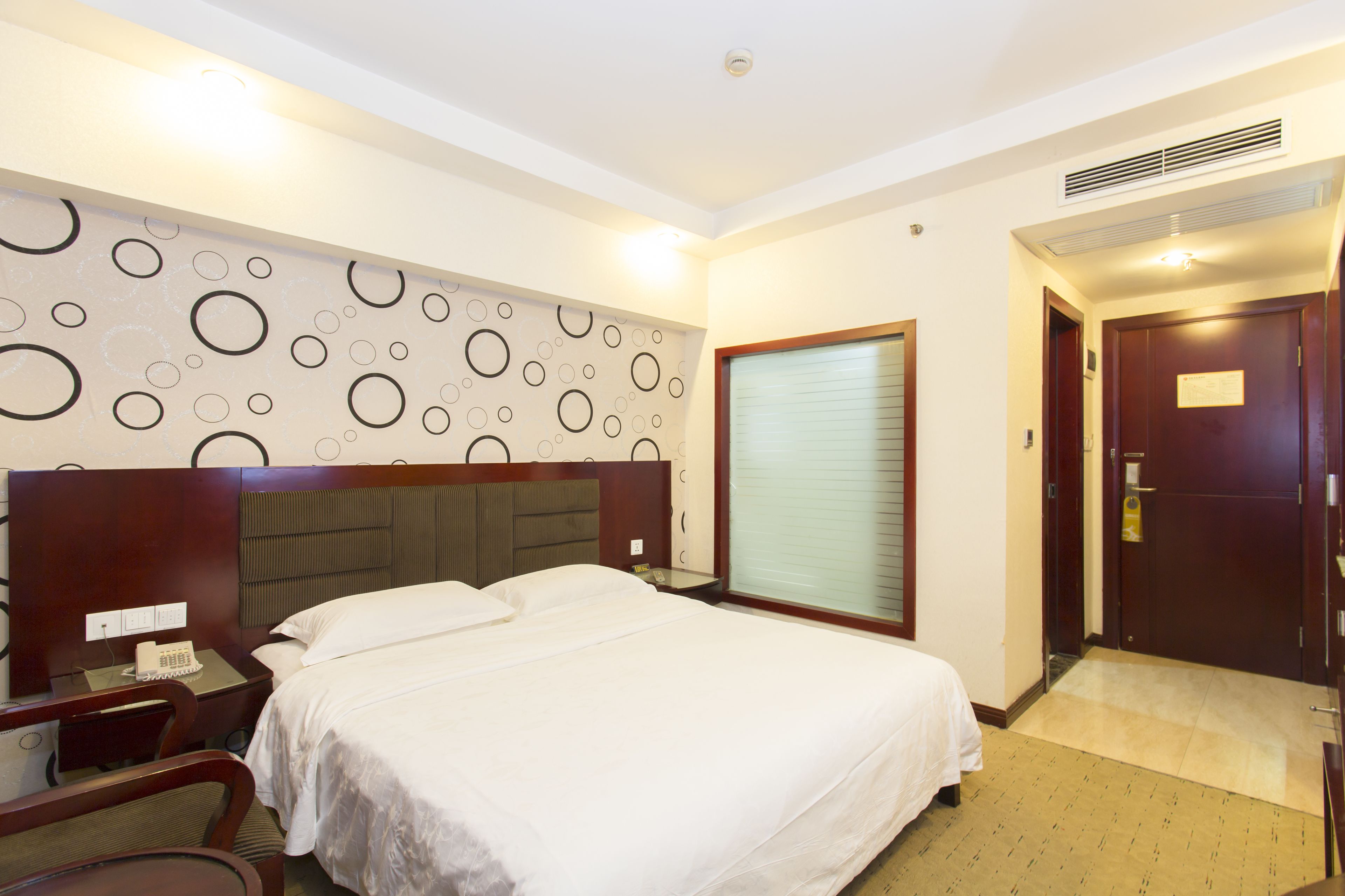 A hotel room with a large bed, a desk with a telephone, a chair, a wall with circular patterns, a window with blinds, and a door with a sign.