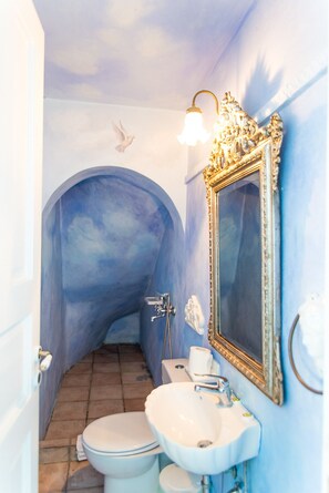 A bathroom with a blue arched doorway, a white toilet, a sink with a mirror, and a light fixture above the mirror. - Cavo Alefkandra 5 (Mykonos)