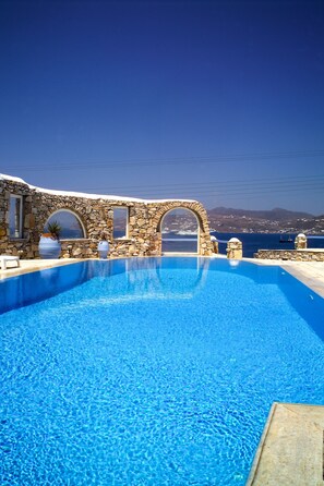 A clear blue pool with stone walls and arched windows, overlooking a coastal landscape. - Cavo Alefkandra 5 (Mykonos)