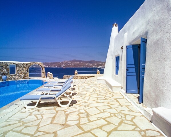 A pool area with lounge chairs, a stone floor, and a view of the sea and distant landscape. - Cavo Alefkandra 5 (Mykonos)
