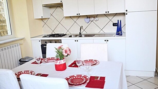 Una cocina compacta con gabinetes blancos, salpicadero de azulejos y un área de comedor para dos personas, con vajilla roja y blanca.