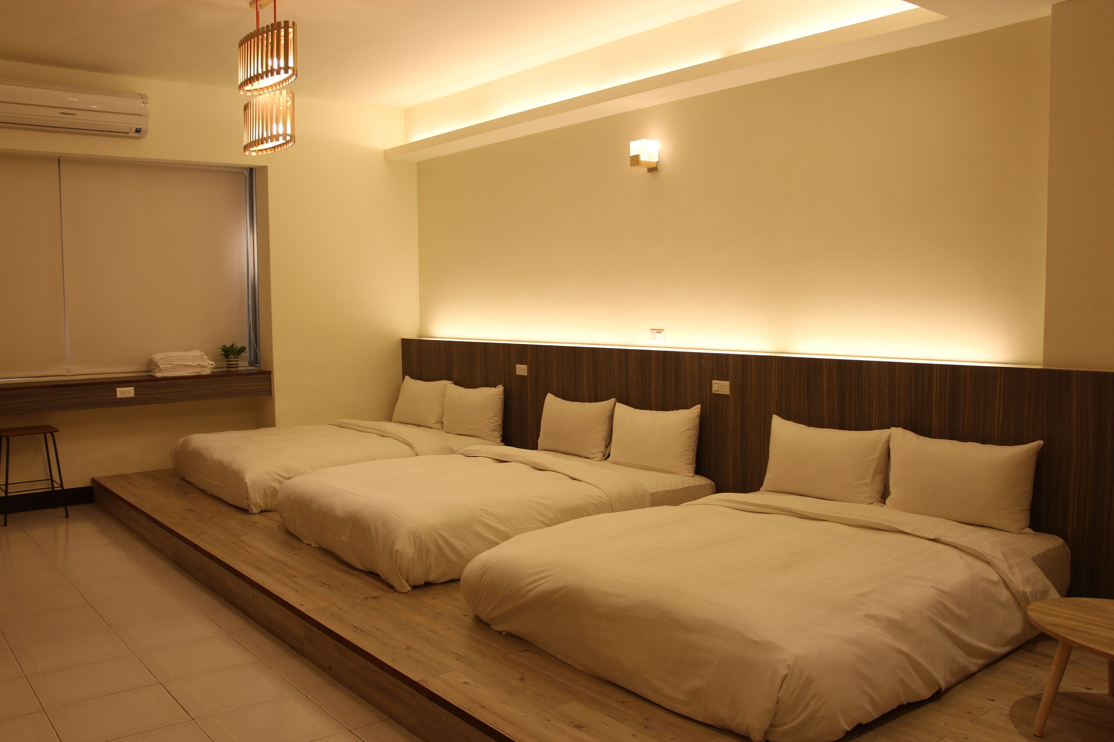 Une chambre d’hôtel avec deux lits, une plateforme en bois, un petit tabouret, une table de chevet et un climatiseur fixé au mur.