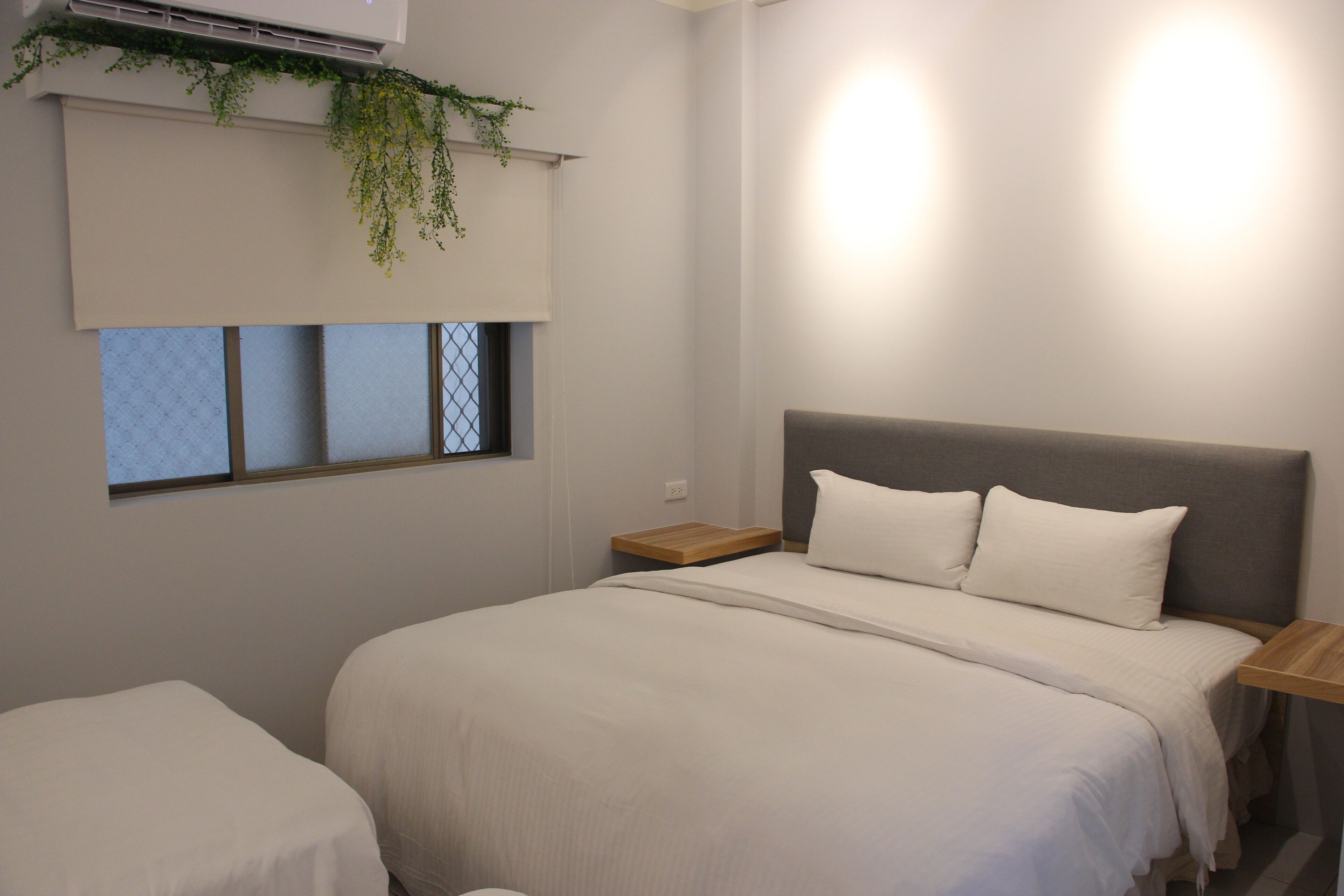 A bedroom with a large bed, a smaller bed, a nightstand, a window with blinds, and an air conditioning unit.