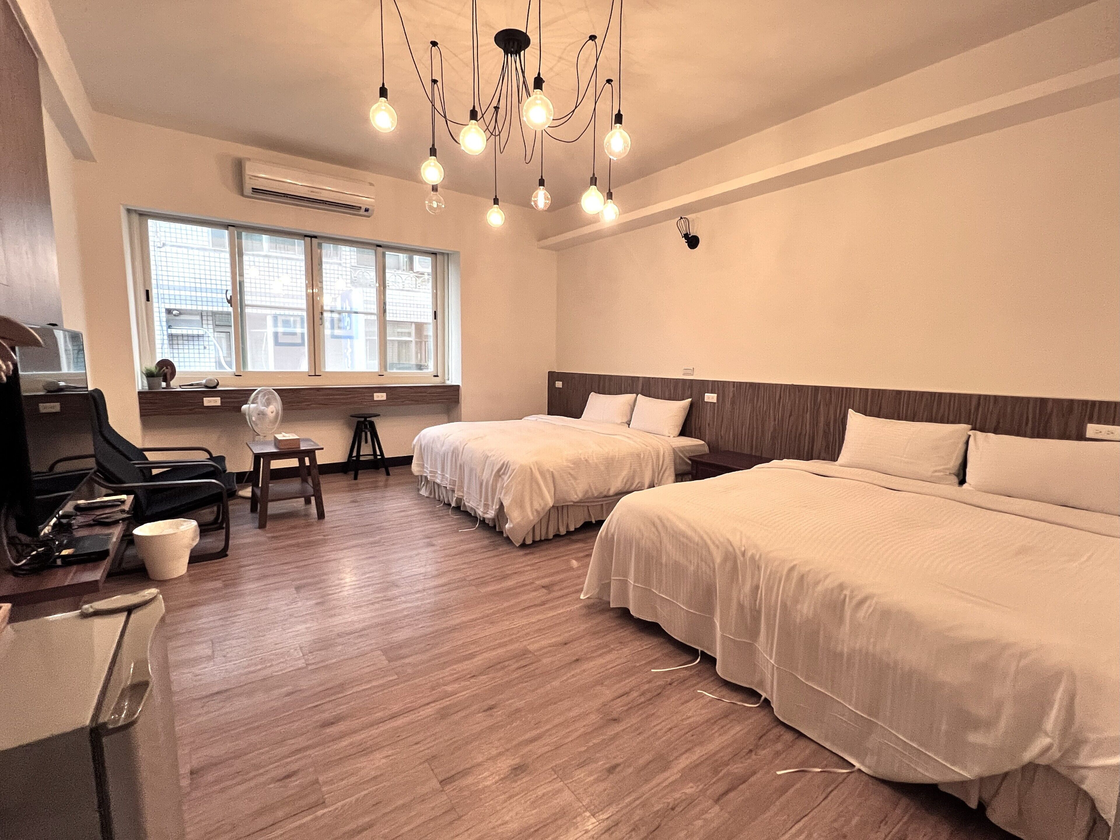 A hotel room with two beds, a desk, a chair, a fan, and a window with a view of buildings.