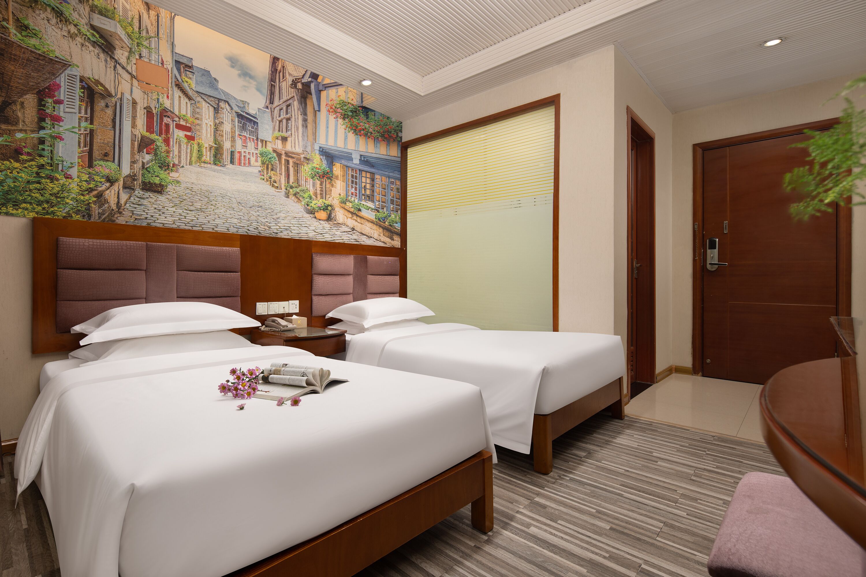 A hotel room with two beds, a large mural of a street scene, a wooden headboard, a bedside table with a lamp, and a large door.