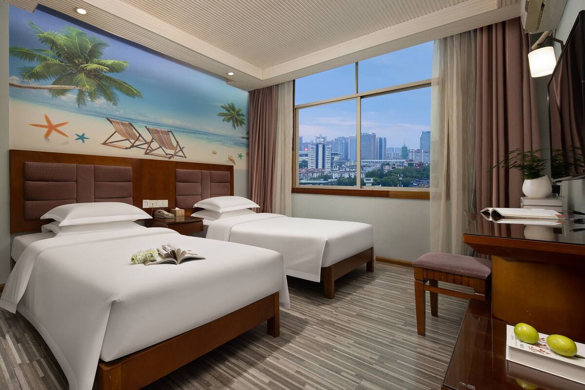 Quarto de hotel com duas camas, uma escrivaninha, uma cadeira, uma janela grande com vista para a cidade e um mural com tema de praia na parede.