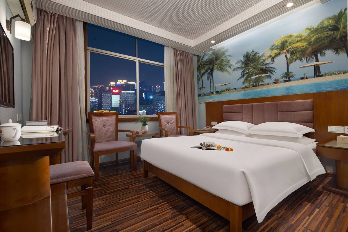 Quarto de hotel com uma cama grande, uma mesa com luminária, uma cadeira e vista para o horizonte noturno.