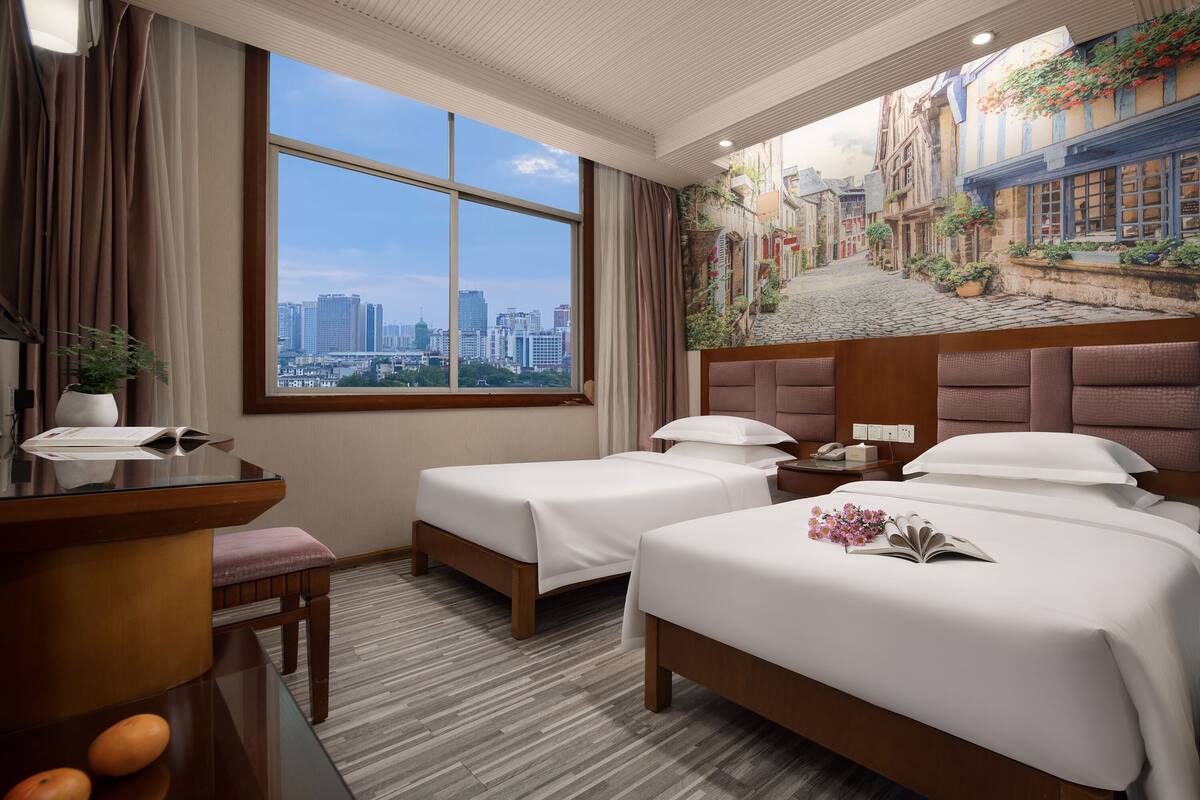 Quarto de hotel com duas camas, uma escrivaninha, uma cadeira, janela com vista para a cidade e um mural de uma cena urbana.