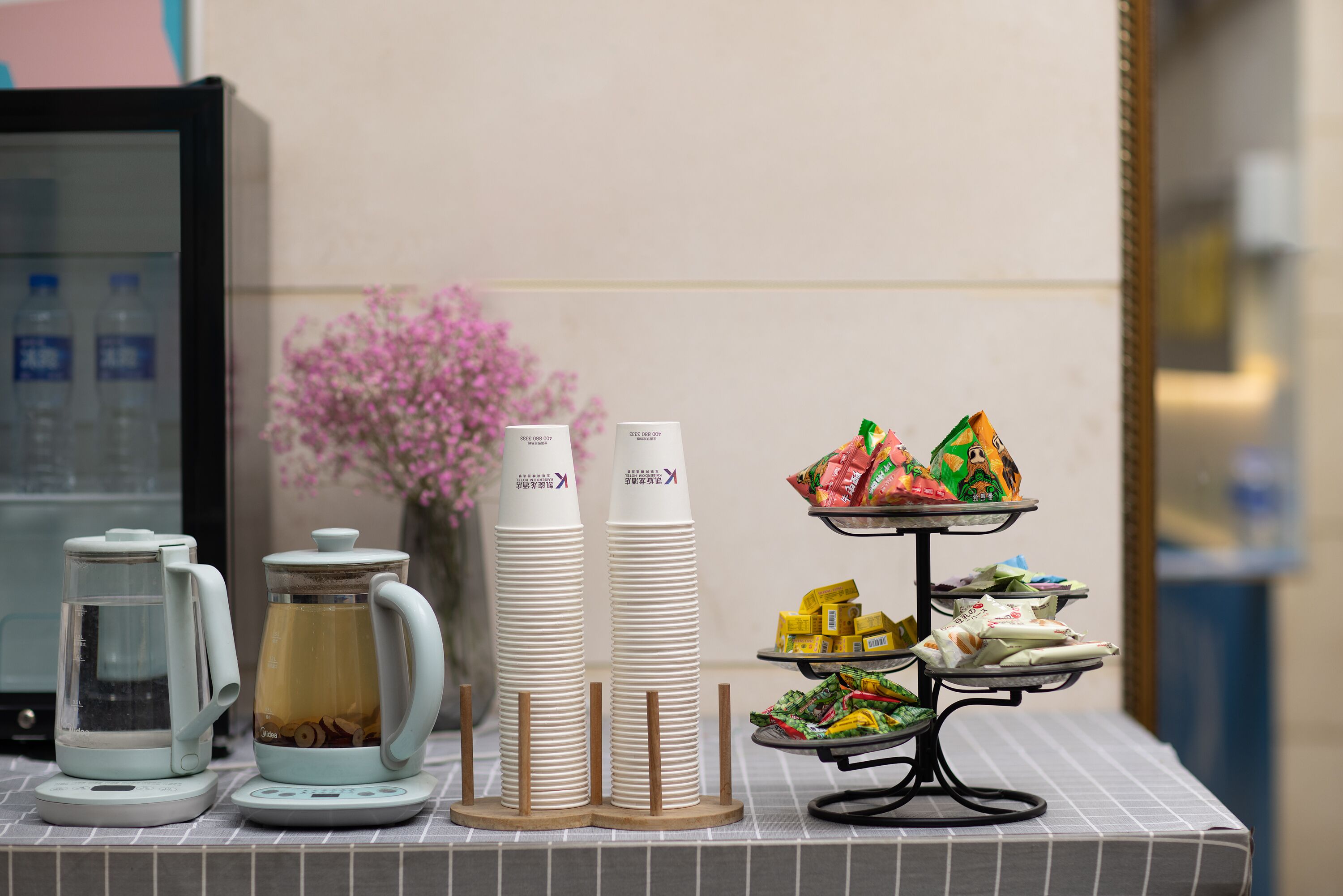 A breakfast setup with a coffee maker, teapot, and stacked cups, along with a tray of assorted pastries and snacks.