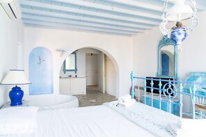 A bedroom with a bed, a white bathtub, a blue lamp, and a bathroom with a sink and mirror. - Cavo Nano (Mykonos)