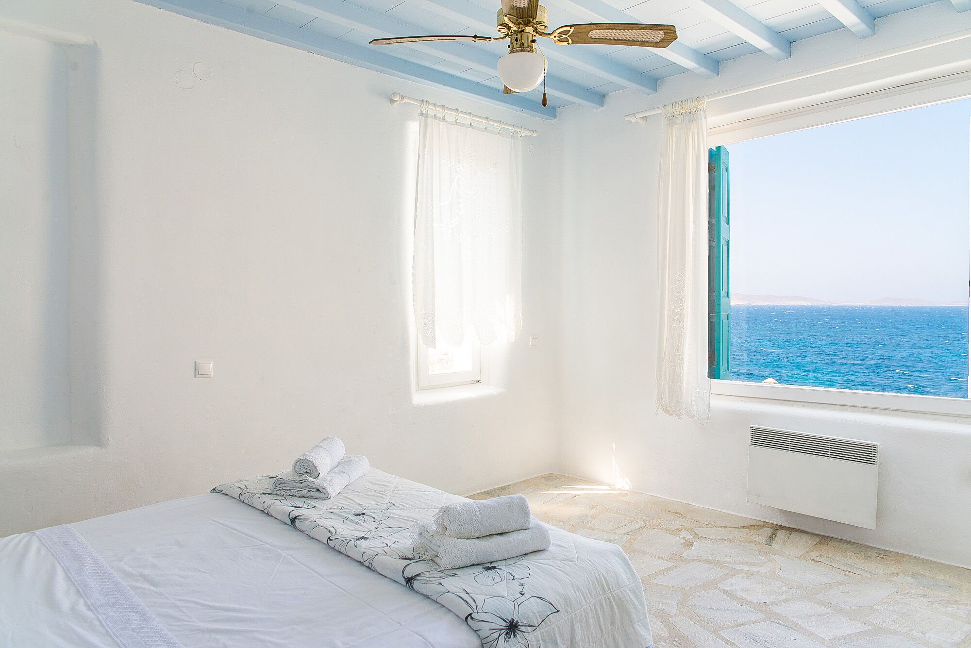 A bedroom with a bed, a ceiling fan, a window with a view of the sea, and a radiator.