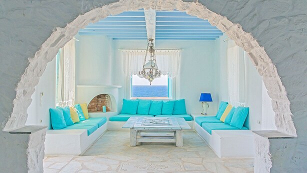 A room with a white archway, a blue sofa set, a chandelier, and a view of the sea. - Cavo Nano (Mykonos)