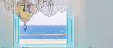 A room with a view of the sea through a window with a white lace curtain.