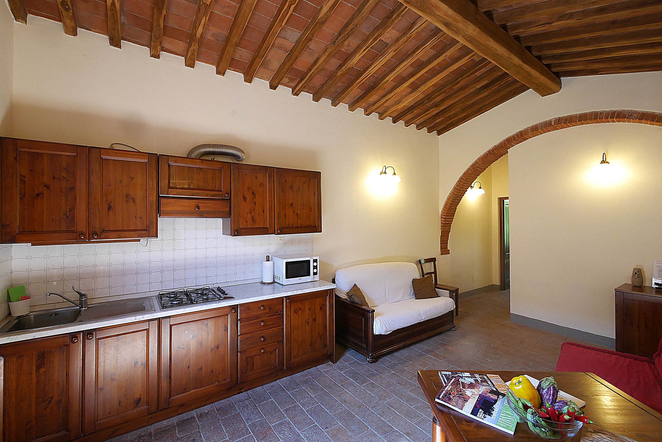Una cocina con armarios de madera, un microondas y un lavabo. Hay un sofá y una mesa con un cuenco de fruta en la sala.