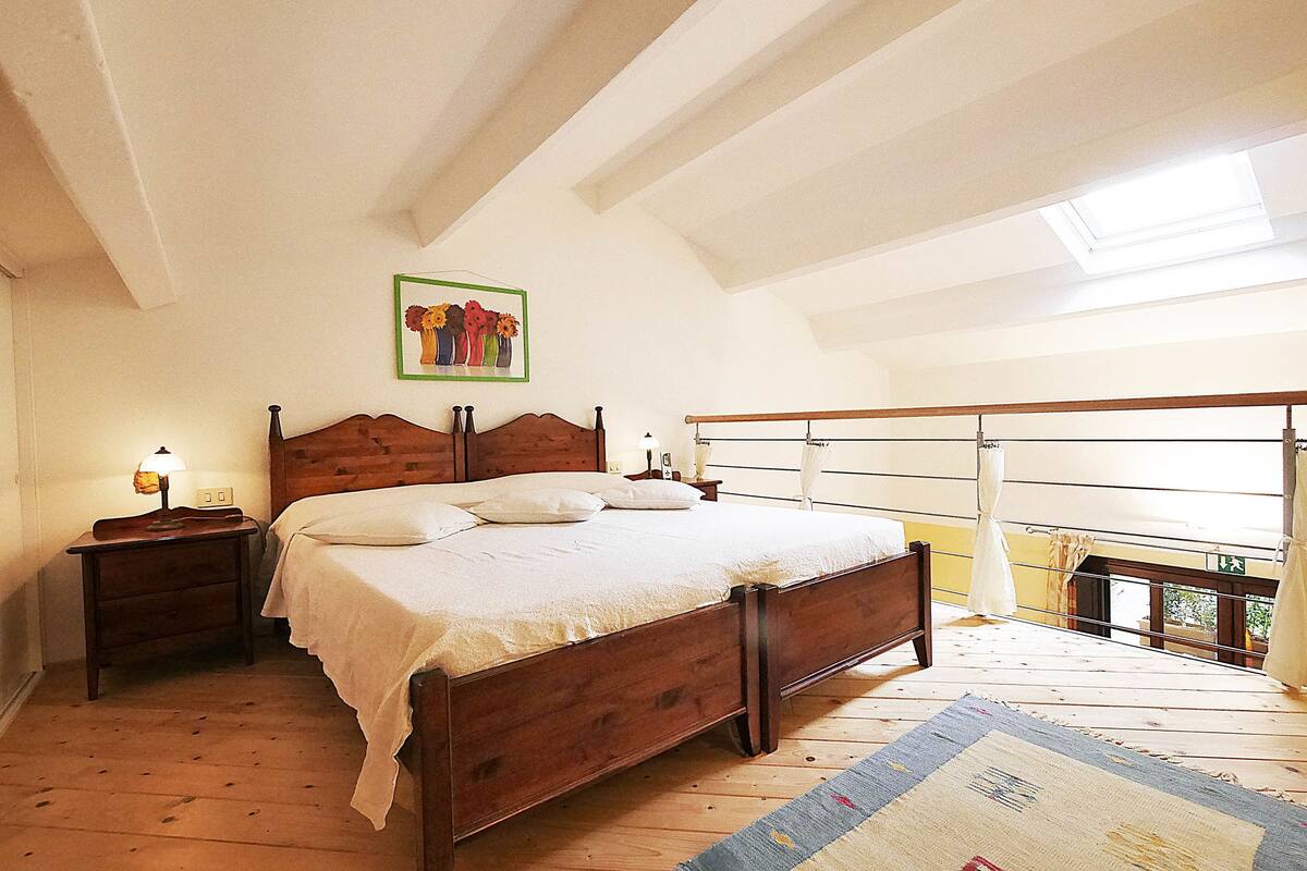 Ein Schlafzimmer mit einem hölzernen Bett, einem Nachttisch mit Lampe, einem Bild an der Wand und einem Balkon mit Geländer.