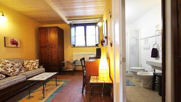 Sala de estar com sofá, mesa e uma cadeira. Há uma janela com cortinas, luminária e porta que dá para um banheiro com vaso sanitário e pia.