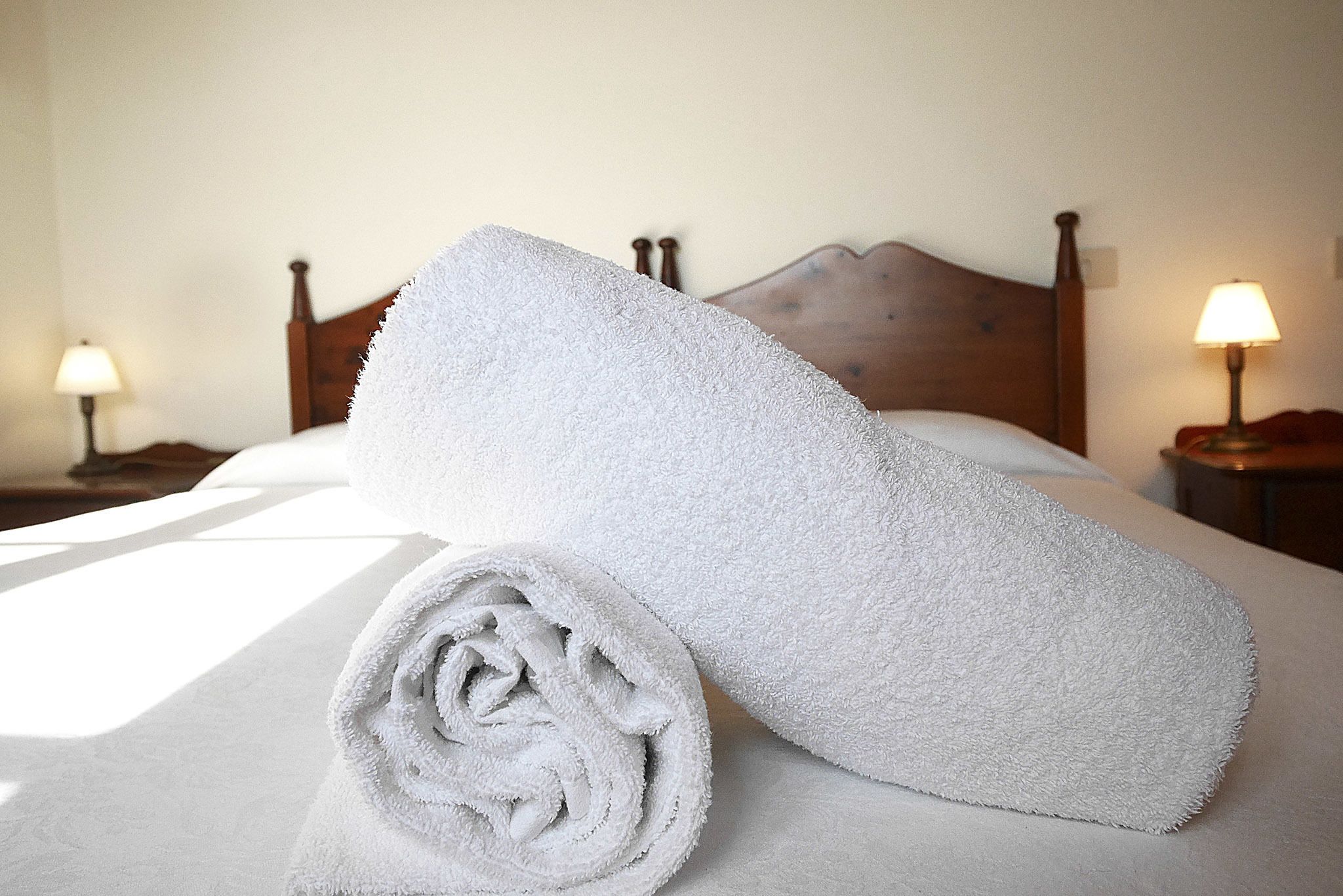 Una cama bien tendida con una toalla enrollada y otra doblada sobre un edredón blanco.