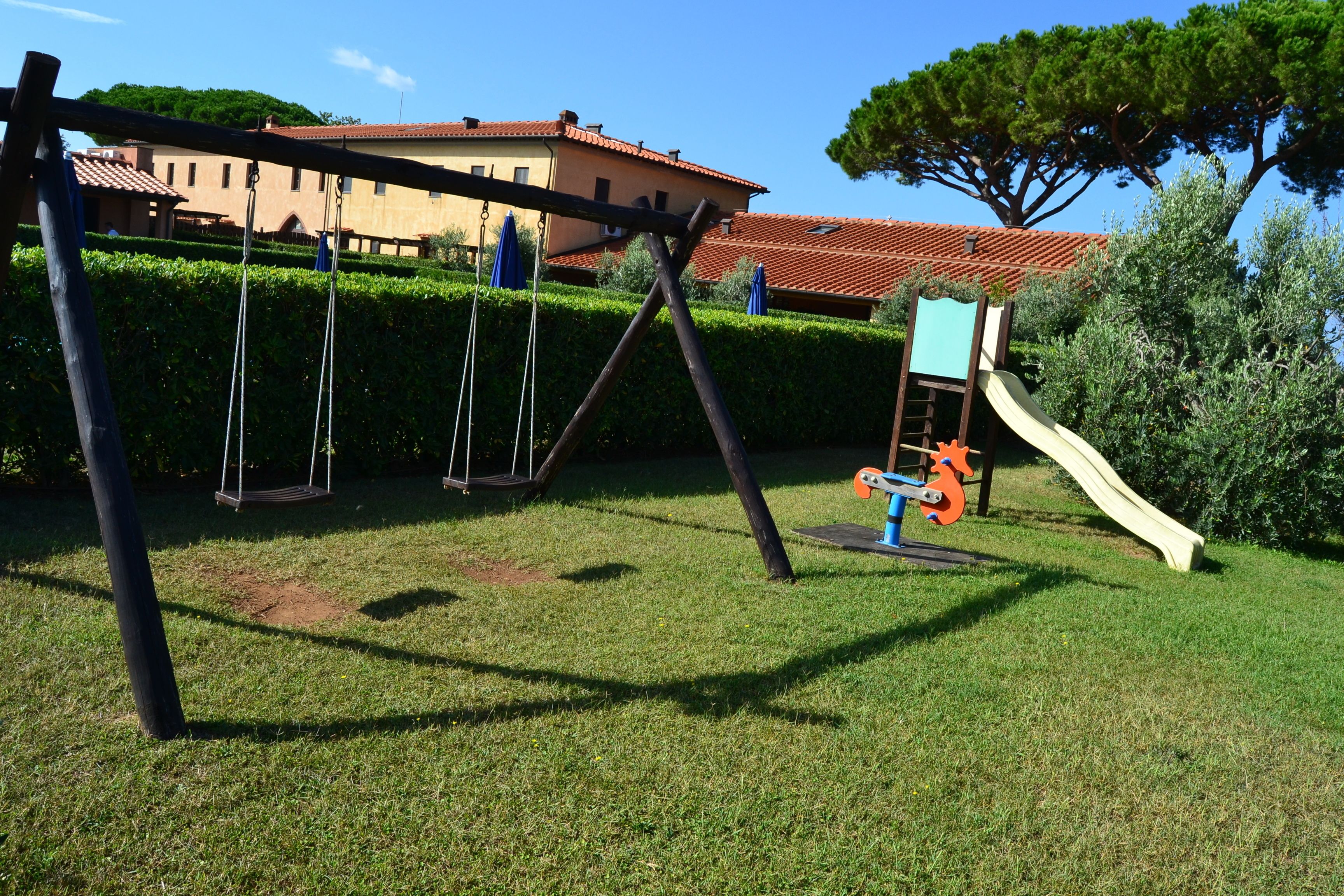 Un parque infantil con columpios y un tobogán, rodeado de césped cuidado y un edificio al fondo.