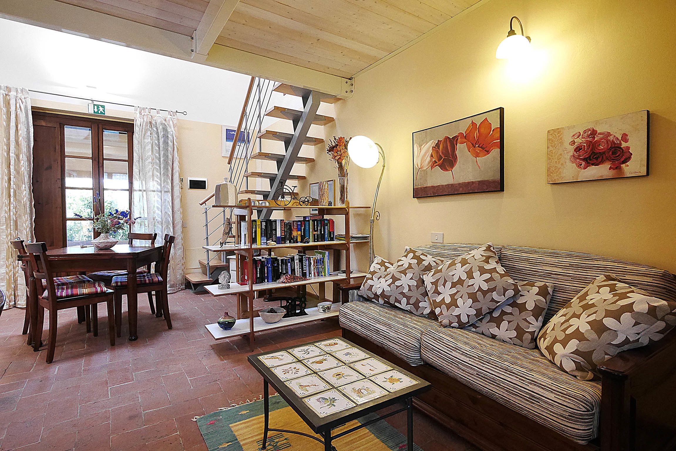 Un salon chaleureux avec un escalier en bois, un canapé agrémenté de coussins à motifs, une table à manger avec des chaises et une bibliothèque remplie de livres.