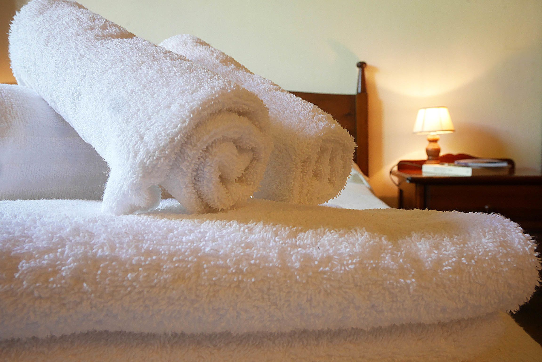A neatly folded white towel on a bed with a wooden headboard and a bedside table with a lamp.