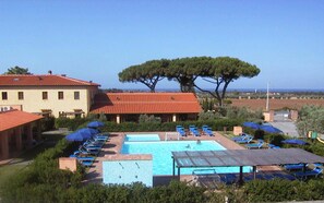 A pool area with multiple sun loungers and umbrellas, a large tree, and a building with a red roof.