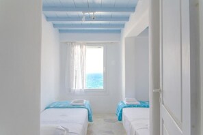 A room with two beds, a window with a view of the sea, and a white door. - Villa Adonis (Mykonos)