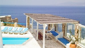 Un área junto a la piscina con sillas reclinables azules, una zona de estar cubierta y vistas al mar.