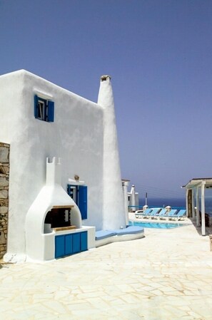 A white building with a blue door and windows, a stone wall, and a pool area with lounge chairs. - Villa Adonis (Mykonos)