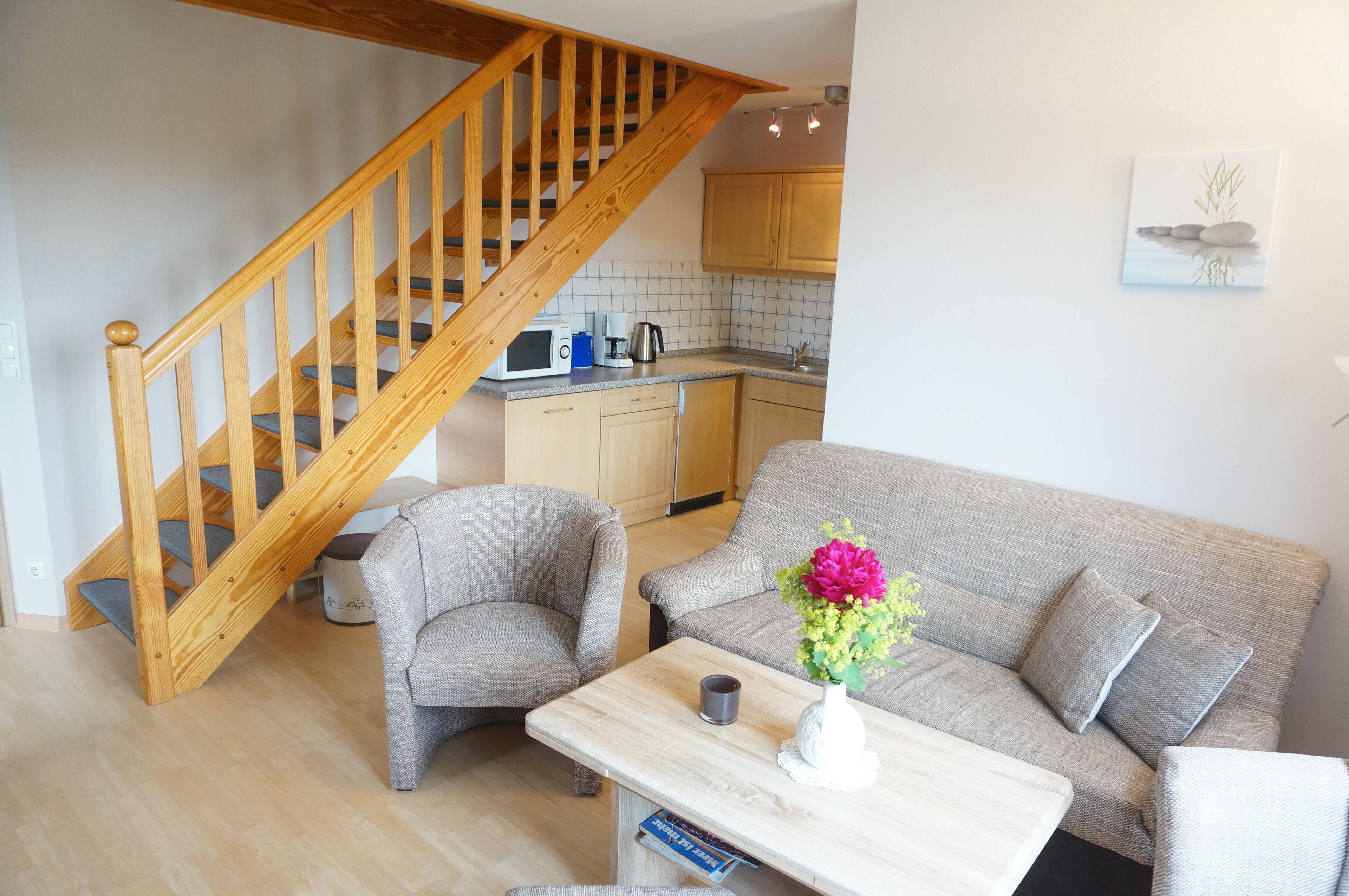 Una sala acogedora con una escalera de madera, un sofá gris, una mesita con un florero y una cocina al fondo.