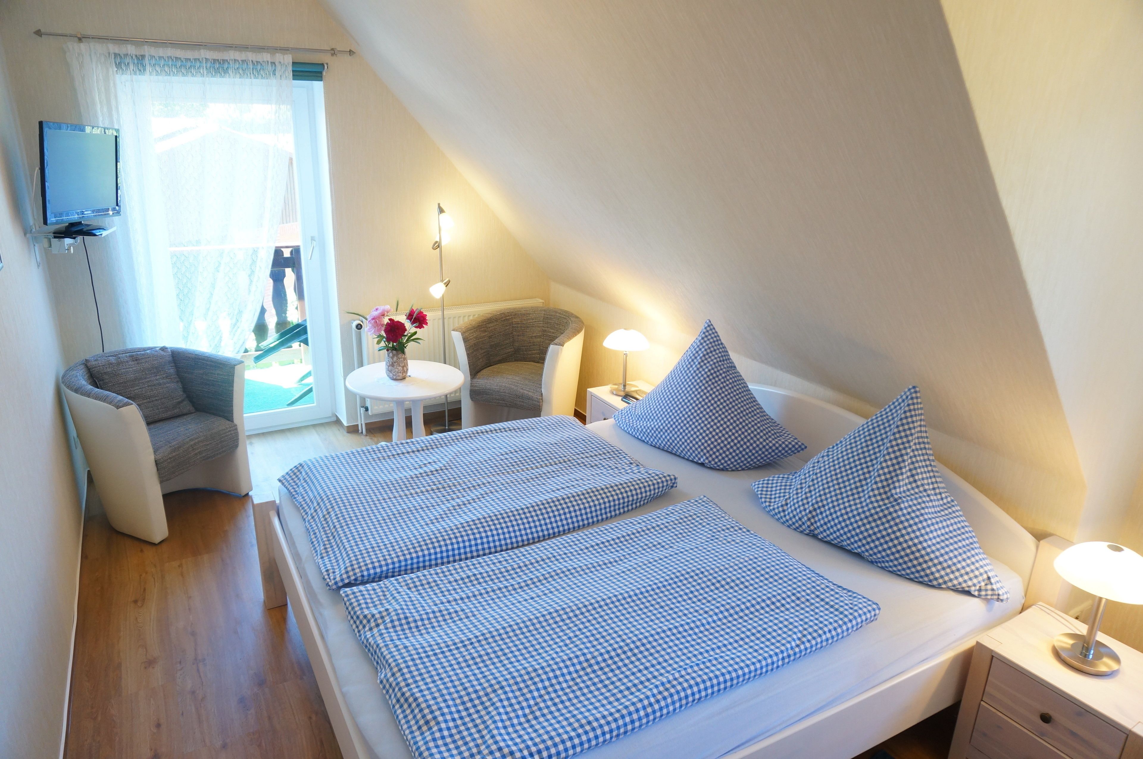 Un dormitorio con cama, una silla, una mesita con flores y un televisor en la pared.