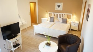 Un dormitorio con cama, mesitas de noche, televisor, una silla y una pequeña mesa con un arreglo floral.