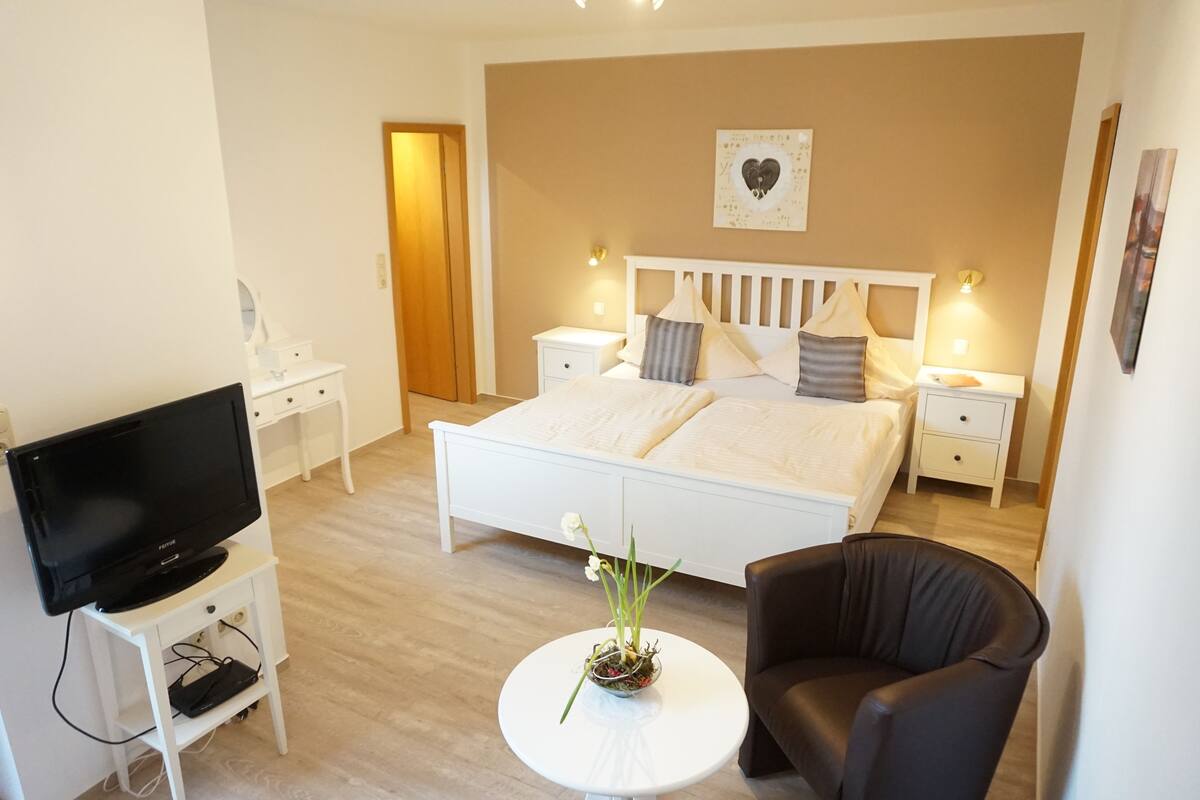 A bedroom with a bed, bedside tables, a TV, a chair, and a small table with a flower arrangement.