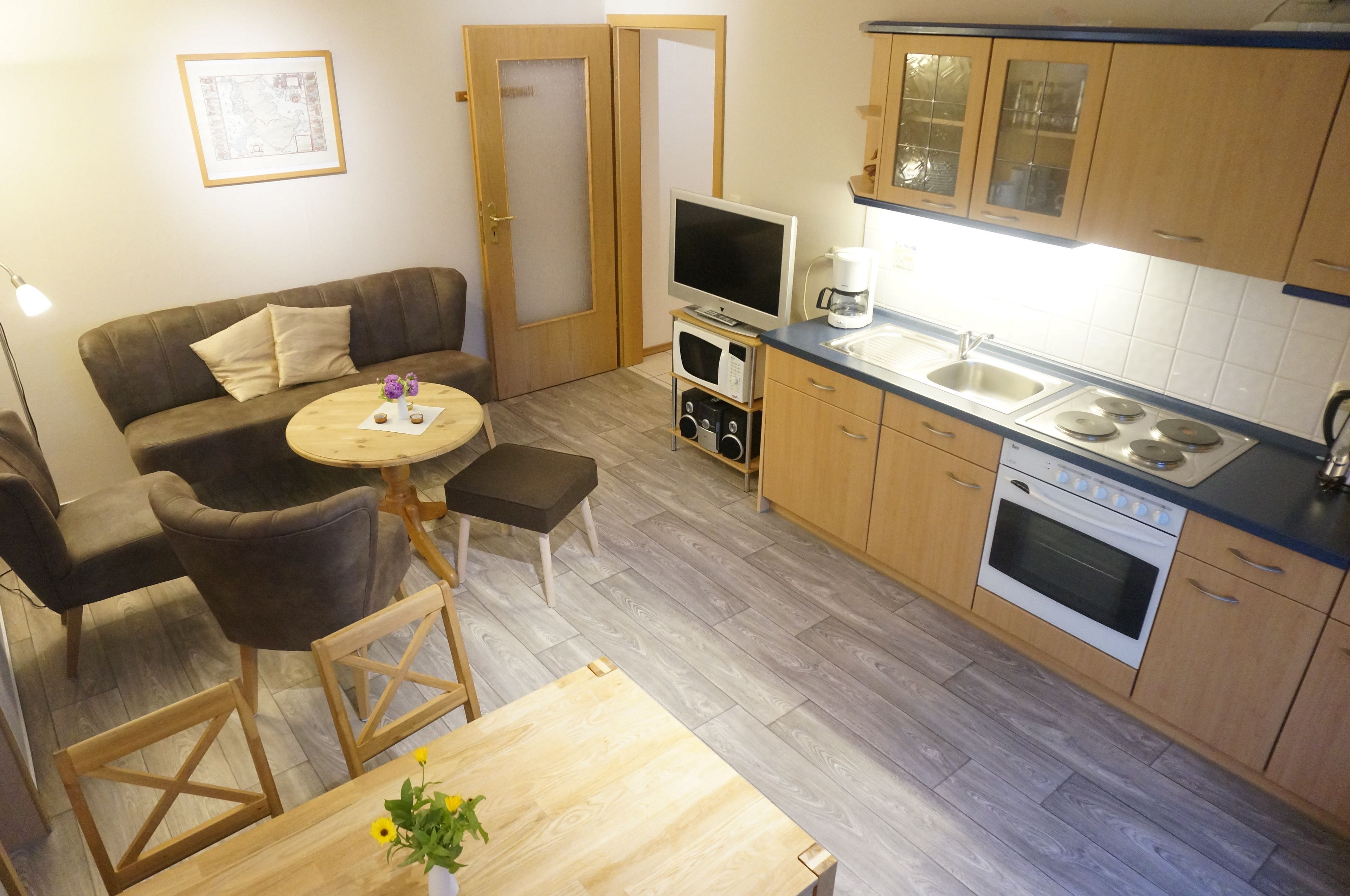 Un espacio habitable compacto con un sofá, una mesita redonda, un televisor, una cafetera y una kitchenette con estufa y lavamanos.