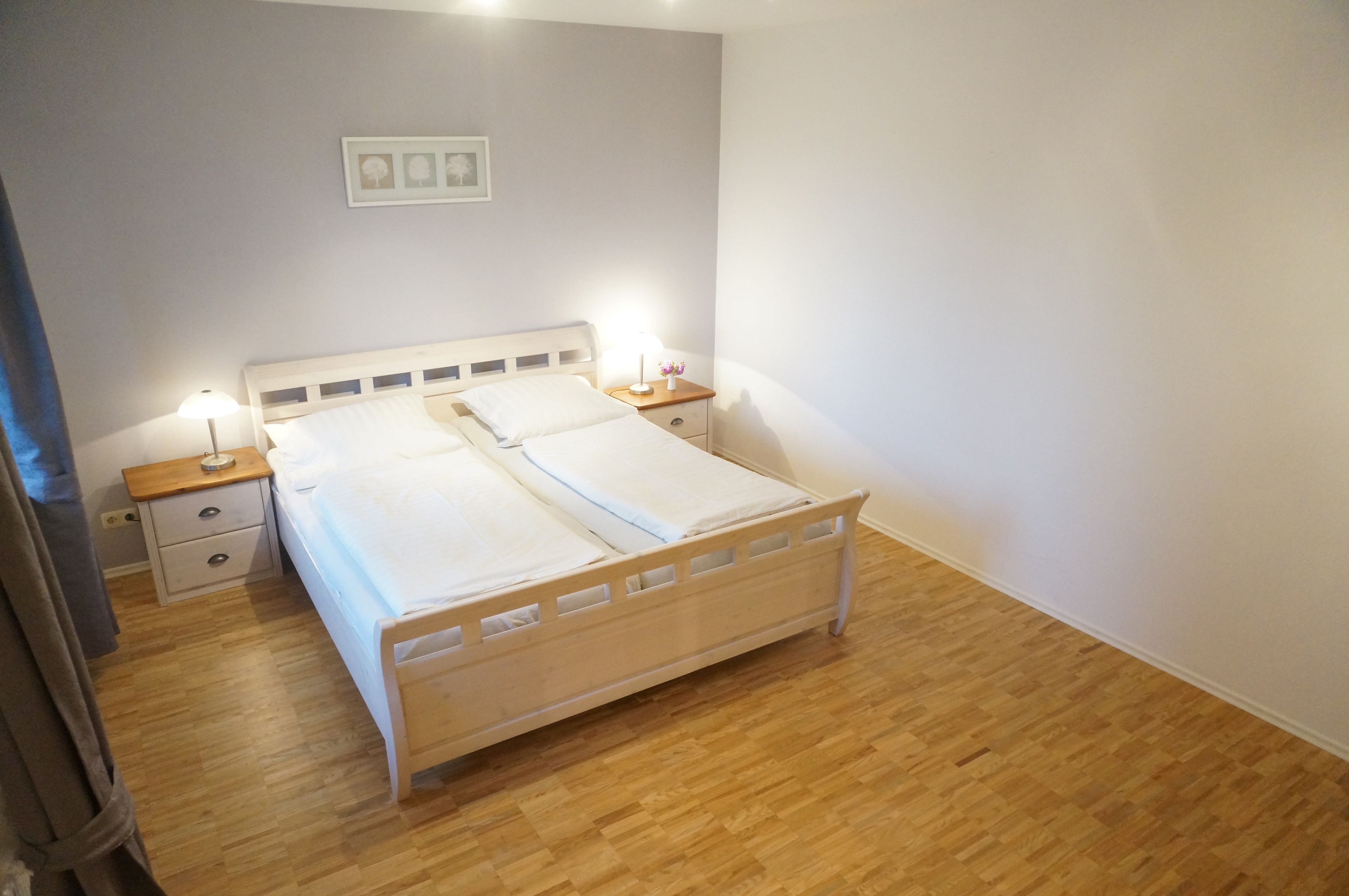 A neatly made bed in a room with a wooden floor, a nightstand with a lamp, and a small drawer.
