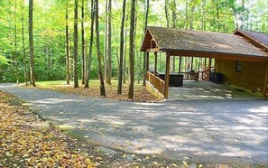 A cabin with a covered porch and a small deck, surrounded by tall trees and a paved driveway. - Cant Wait to Hibernate (Sevierville)