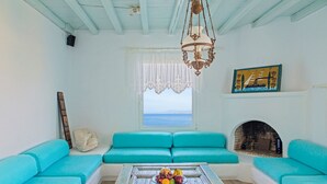 A room with turquoise sofas, a wooden coffee table with fruits, a white fireplace, and a view of the sea. - Cavo Delos (Mykonos)