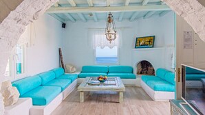 A living room with a blue sofa set, a coffee table with fruit, a chandelier, and a view of the sea through a window. - Cavo Delos (Mykonos)