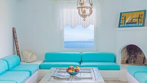 A room with a blue sofa, a wooden coffee table with fruits, a hanging light fixture, and a view of the sea through a window. - Cavo Delos (Mykonos)