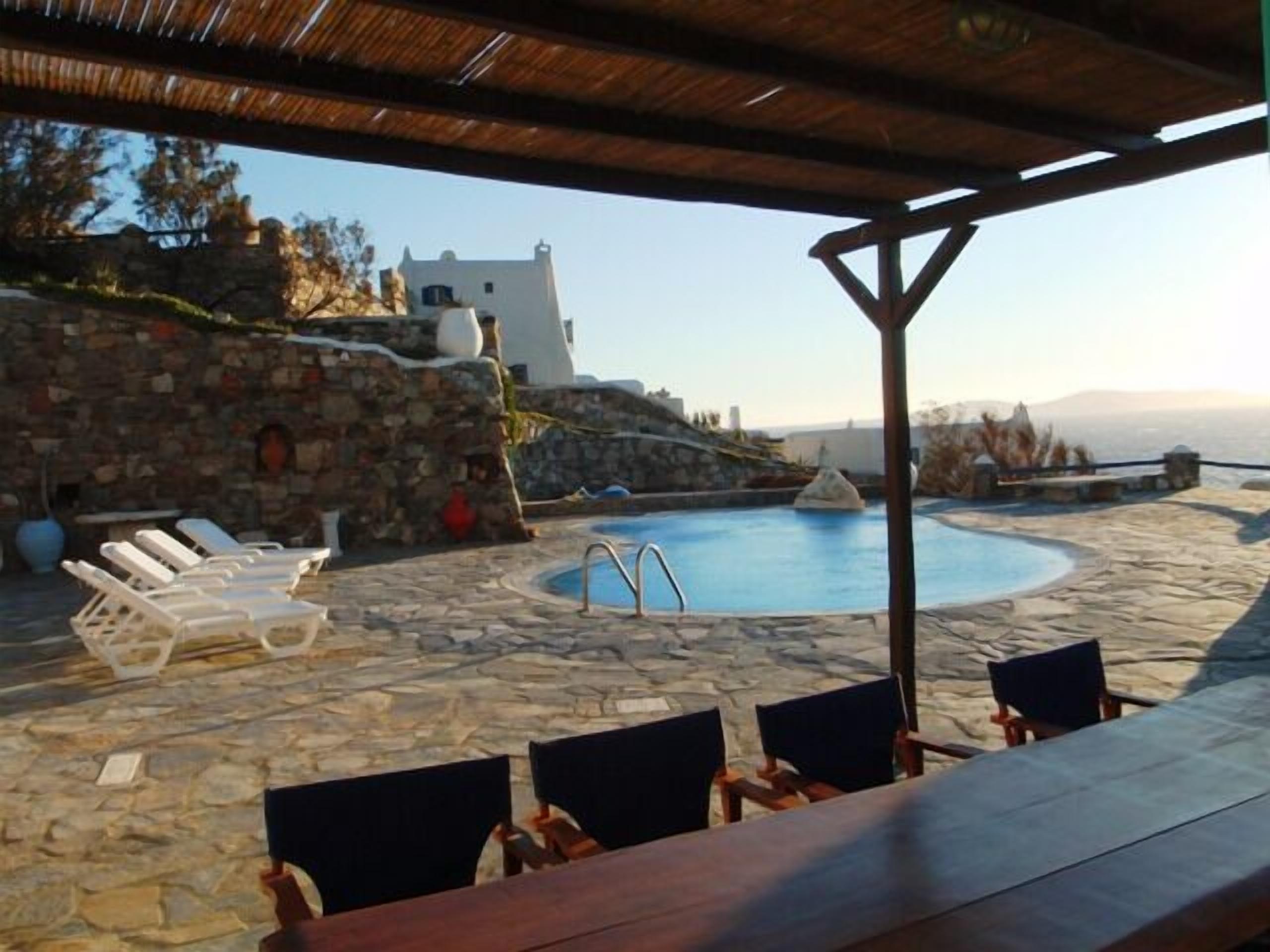 Uma piscina de pedra com espreguiçadeiras e uma mesa, cercada por muros de pedra e vista para o mar.