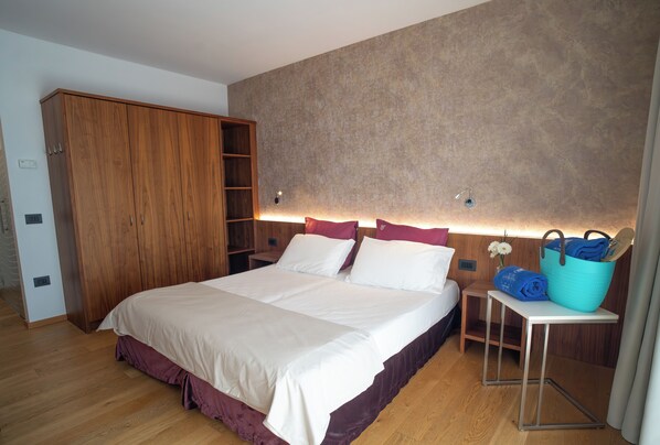 Un dormitorio con una cama grande, un armario de madera, una mesita de noche con una lámpara y una pequeña mesa con una cesta azul.