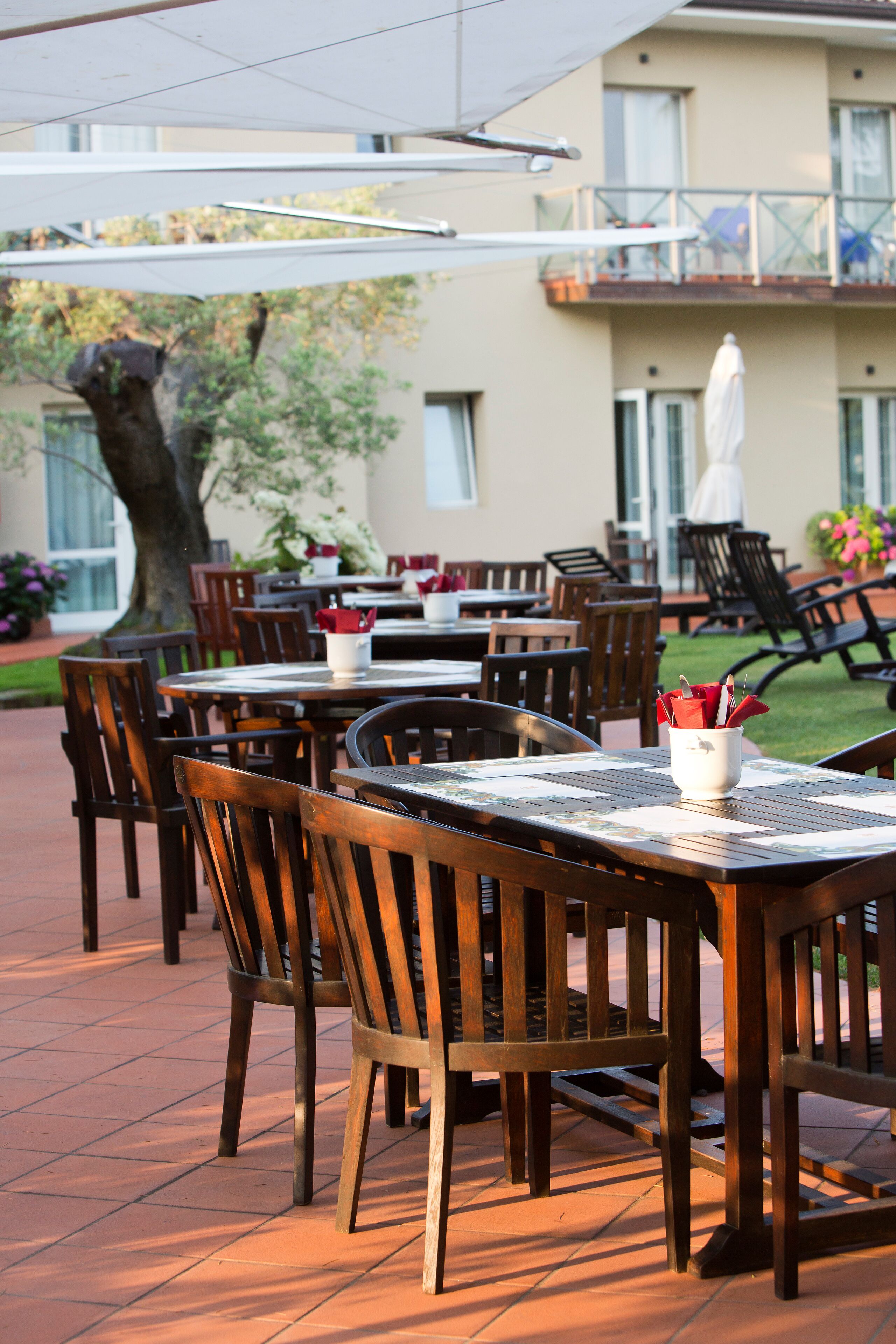 An outdoor patio with wooden tables and chairs, a white umbrella, and a building in the background.